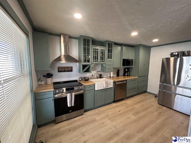 kitchen with sink, wooden counters, green cabinets, stainless steel appliances, and wall chimney exhaust hood