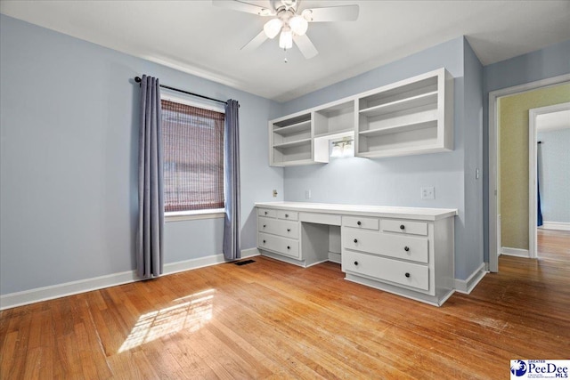 unfurnished office featuring light wood-type flooring, ceiling fan, baseboards, and built in desk