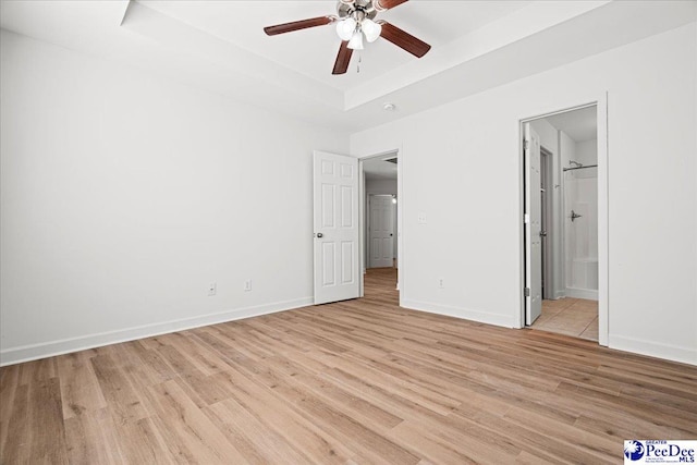 unfurnished bedroom with baseboards, a raised ceiling, a ceiling fan, ensuite bath, and light wood-style flooring