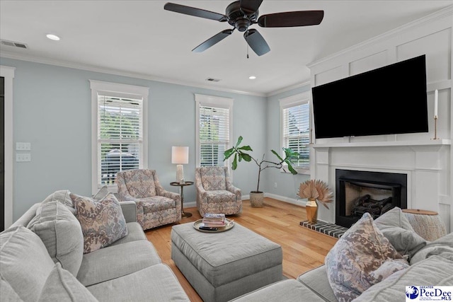 living room with healthy amount of natural light, crown molding, a fireplace with raised hearth, a ceiling fan, and wood finished floors