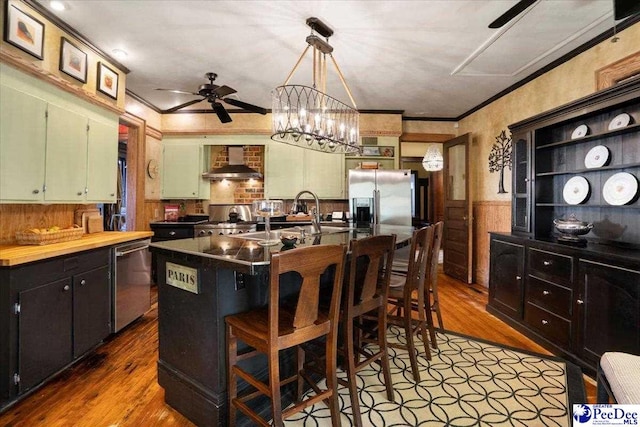 kitchen with sink, a kitchen island with sink, stainless steel appliances, crown molding, and wall chimney exhaust hood