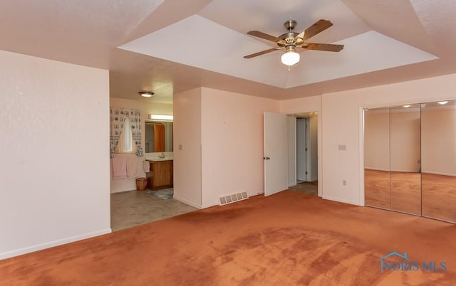 unfurnished bedroom with ensuite bath, ceiling fan, a raised ceiling, light colored carpet, and a closet