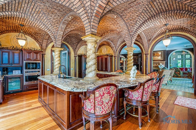 kitchen with light stone counters, a center island with sink, stainless steel appliances, sink, and decorative columns