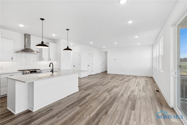 kitchen featuring white cabinets, hanging light fixtures, an island with sink, wall chimney range hood, and light hardwood / wood-style flooring