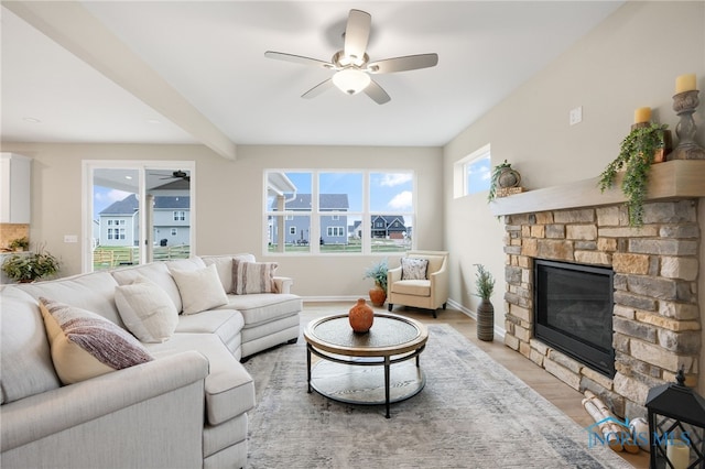 living room with beamed ceiling, light wood-type flooring, a stone fireplace, and ceiling fan