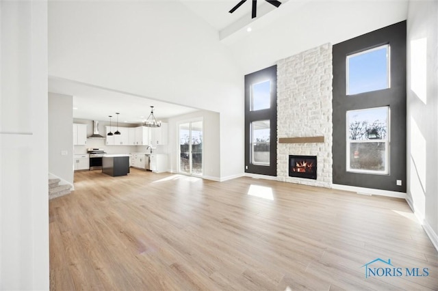 unfurnished living room with ceiling fan with notable chandelier, a high ceiling, a stone fireplace, light wood finished floors, and baseboards