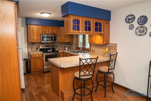 kitchen with glass insert cabinets, brown cabinets, a peninsula, stainless steel appliances, and a sink