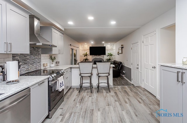 kitchen with kitchen peninsula, light stone countertops, stainless steel appliances, light wood-type flooring, and wall chimney range hood