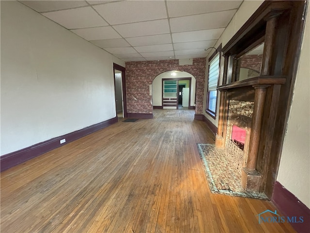 unfurnished living room featuring baseboards, arched walkways, brick wall, dark wood-type flooring, and a paneled ceiling