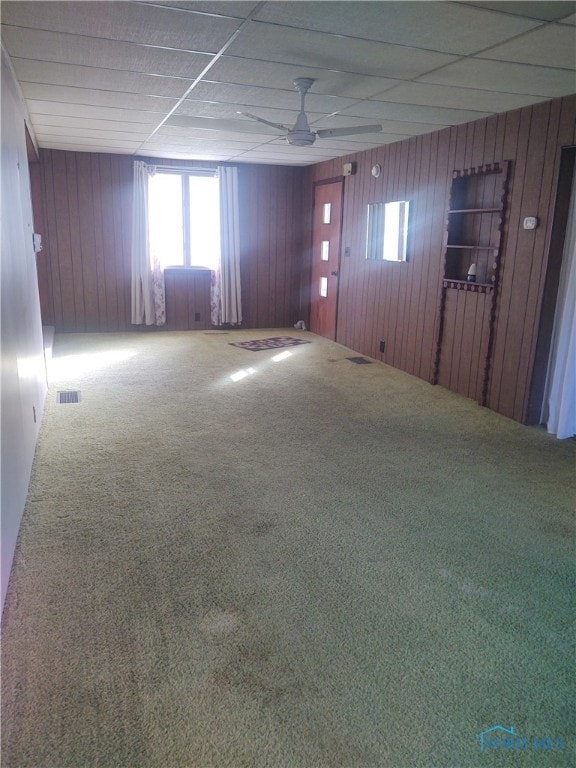 carpeted empty room featuring a drop ceiling, wooden walls, and ceiling fan