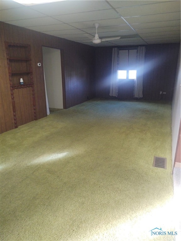 empty room with carpet floors, wood walls, a drop ceiling, and ceiling fan