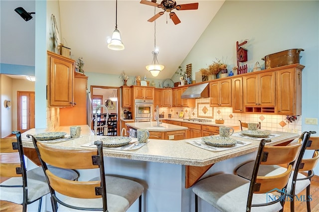 kitchen with kitchen peninsula, appliances with stainless steel finishes, wall chimney exhaust hood, light hardwood / wood-style flooring, and high vaulted ceiling