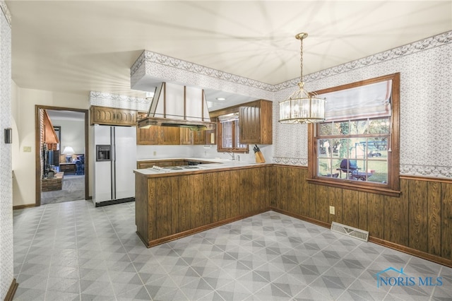 kitchen featuring white fridge with ice dispenser, island range hood, kitchen peninsula, pendant lighting, and wooden walls