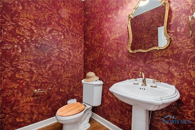 half bathroom with wallpapered walls, wood finished floors, toilet, and baseboards