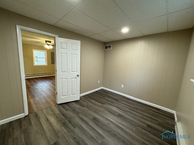 empty room with a baseboard heating unit, ceiling fan, dark hardwood / wood-style flooring, wood walls, and a drop ceiling