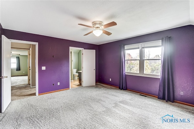 unfurnished bedroom featuring ensuite bathroom, light colored carpet, multiple windows, and ceiling fan