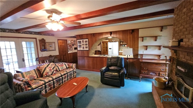 carpeted living room featuring wood walls, ceiling fan, beam ceiling, a fireplace, and french doors