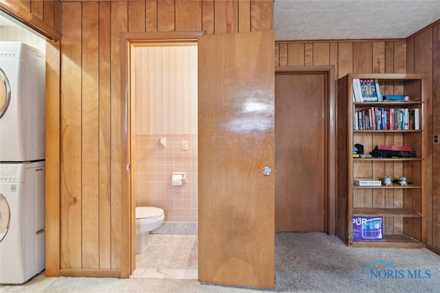 bathroom with wooden walls, stacked washer / dryer, tile walls, tile patterned flooring, and toilet