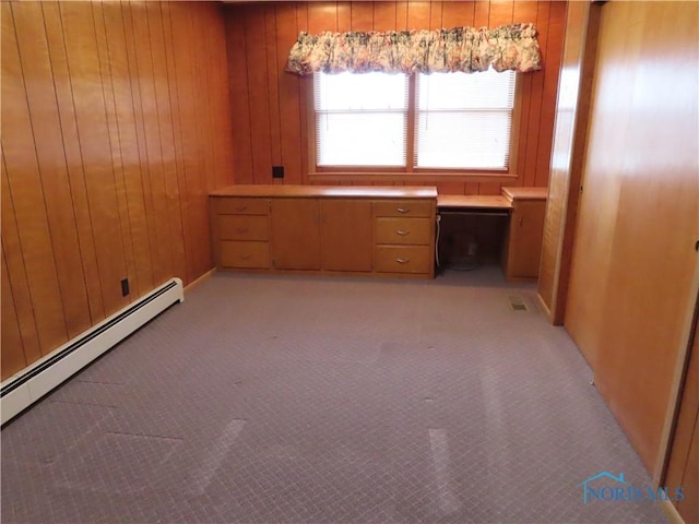 unfurnished office featuring wood walls, light colored carpet, built in desk, and a baseboard heating unit