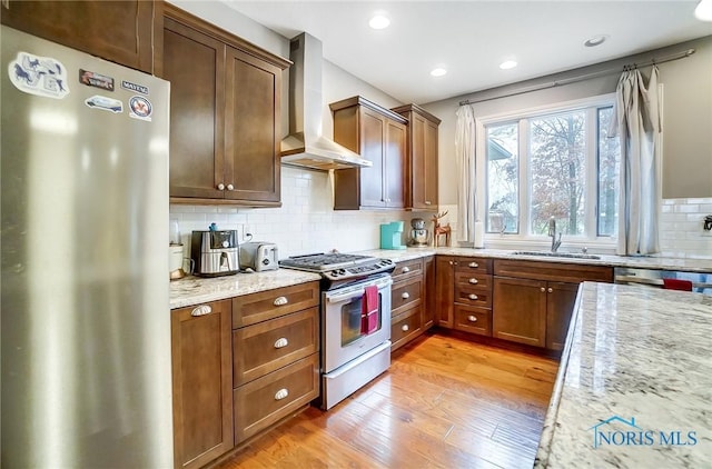 kitchen with wall chimney range hood, sink, light wood-type flooring, light stone counters, and stainless steel appliances