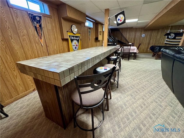 bar with tile counters, light colored carpet, wood walls, and a drop ceiling