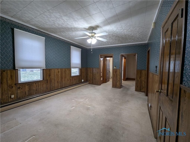 carpeted spare room with baseboard heating, ceiling fan, wood walls, and ornamental molding