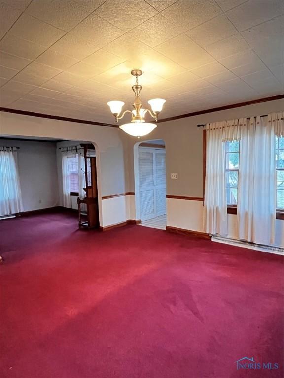 carpeted empty room with a notable chandelier, crown molding, and a baseboard heating unit