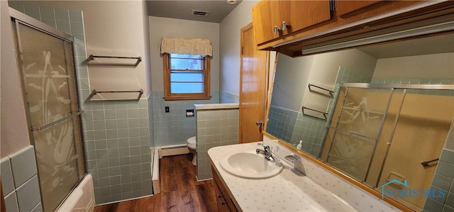 full bathroom with bath / shower combo with glass door, baseboard heating, wood-type flooring, tile walls, and toilet