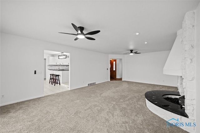 unfurnished living room with ceiling fan, a stone fireplace, and light carpet