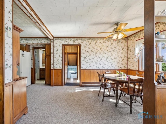 dining area featuring wood walls, a baseboard heating unit, crown molding, ceiling fan, and light colored carpet