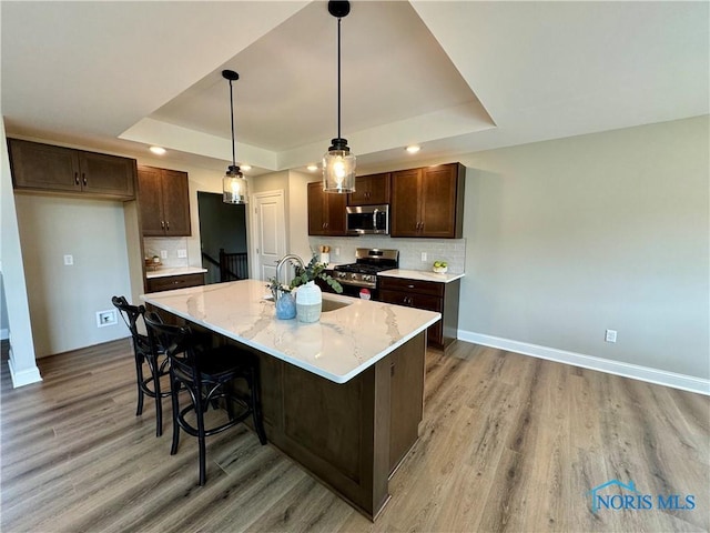 kitchen with hanging light fixtures, a raised ceiling, sink, appliances with stainless steel finishes, and a center island with sink