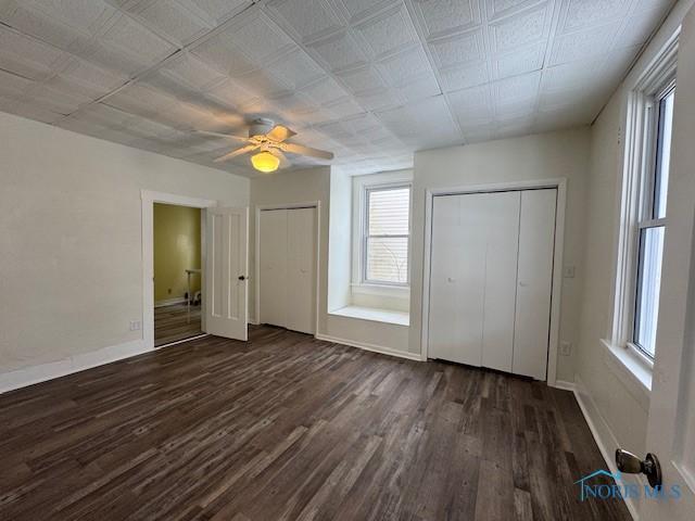 unfurnished bedroom with multiple windows, ceiling fan, dark wood-type flooring, and multiple closets