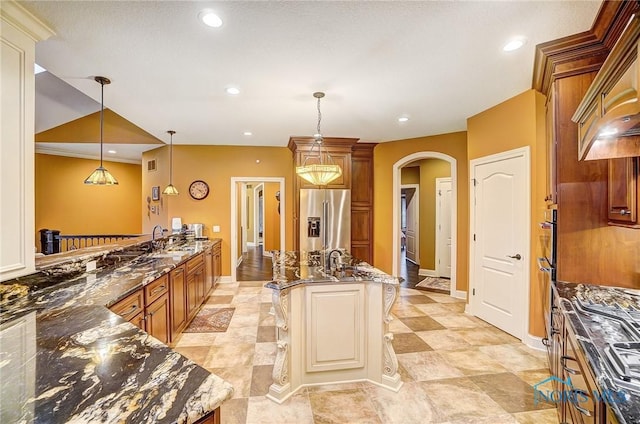 kitchen with a kitchen bar, high end fridge, a kitchen island with sink, decorative light fixtures, and dark stone countertops