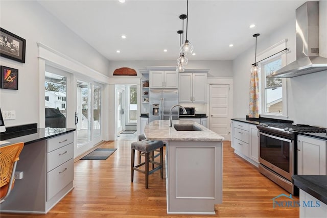 kitchen with sink, wall chimney range hood, decorative light fixtures, a kitchen island with sink, and appliances with stainless steel finishes