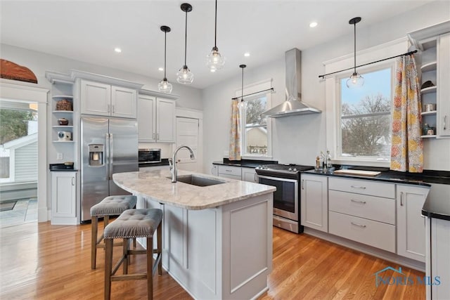 kitchen featuring sink, wall chimney exhaust hood, stainless steel appliances, decorative light fixtures, and a center island with sink