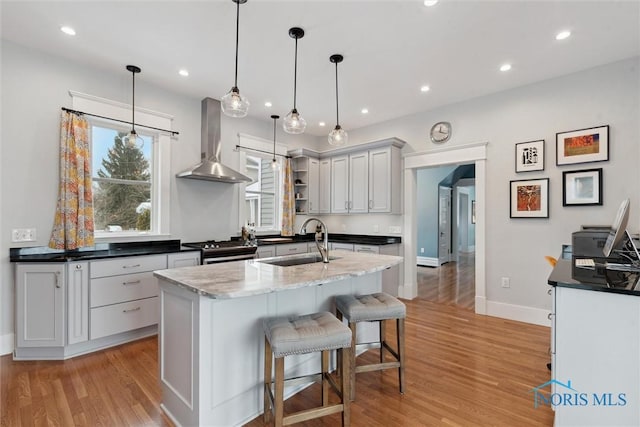 kitchen featuring sink, wall chimney range hood, decorative light fixtures, a center island with sink, and light hardwood / wood-style flooring