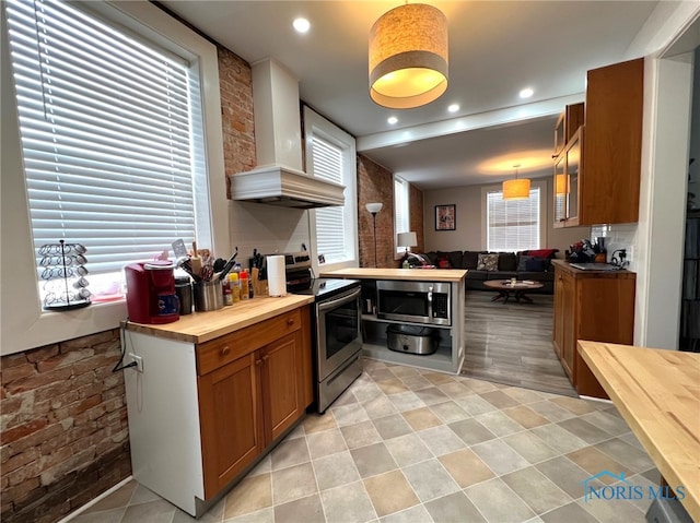 kitchen with pendant lighting, butcher block countertops, appliances with stainless steel finishes, brick wall, and wall chimney exhaust hood