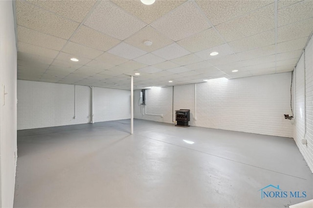 basement featuring brick wall, a drop ceiling, and a wood stove