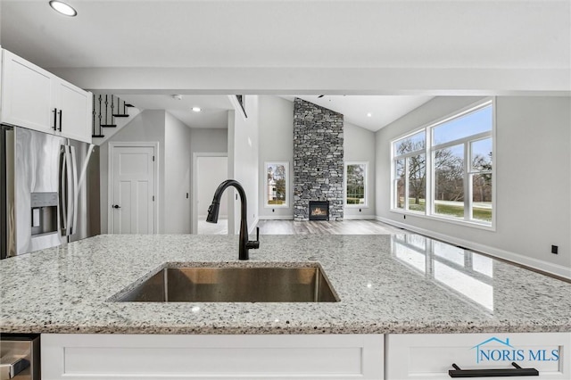 kitchen featuring sink, stainless steel refrigerator with ice dispenser, vaulted ceiling, a fireplace, and light stone counters