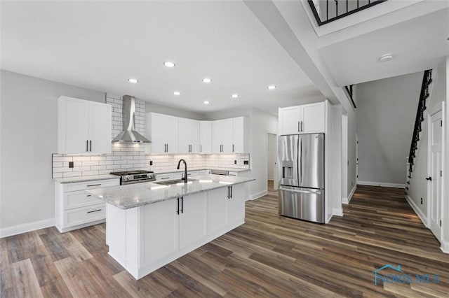 kitchen with white cabinets, wall chimney exhaust hood, light stone countertops, an island with sink, and appliances with stainless steel finishes