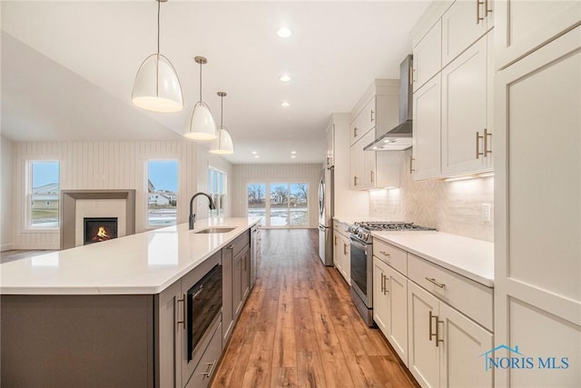 kitchen featuring a large island with sink, sink, hanging light fixtures, appliances with stainless steel finishes, and wall chimney exhaust hood