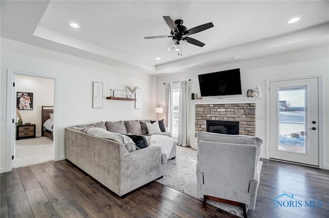 living room with ceiling fan, a stone fireplace, dark wood-type flooring, and a tray ceiling