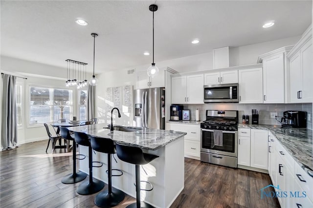 kitchen with white cabinetry, hanging light fixtures, stainless steel appliances, backsplash, and a kitchen island with sink