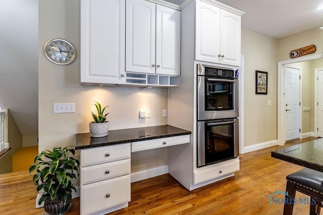 kitchen featuring built in desk, double oven, white cabinetry, dark stone counters, and light wood-type flooring