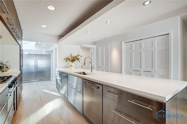 kitchen with sink, a large island with sink, stainless steel appliances, dark brown cabinets, and light hardwood / wood-style flooring