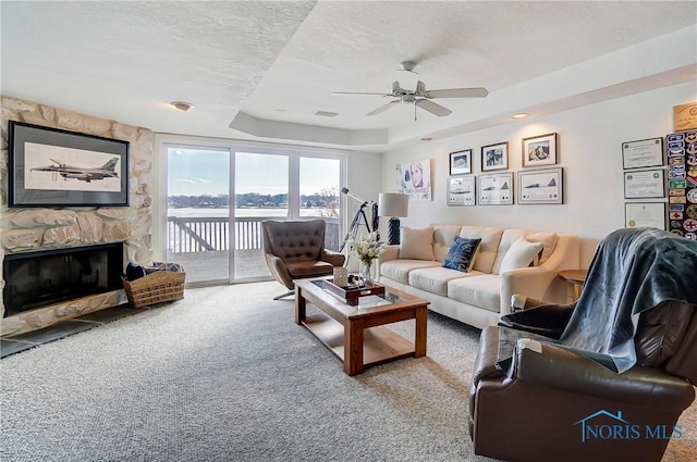 carpeted living room featuring a tray ceiling, ceiling fan, a stone fireplace, and a textured ceiling