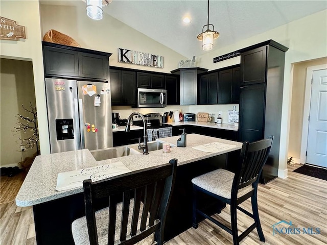 kitchen featuring vaulted ceiling, hanging light fixtures, stainless steel appliances, light stone countertops, and a kitchen island with sink