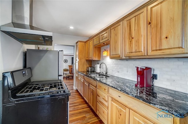 kitchen with gas stove, stainless steel refrigerator, sink, dark stone counters, and wall chimney exhaust hood