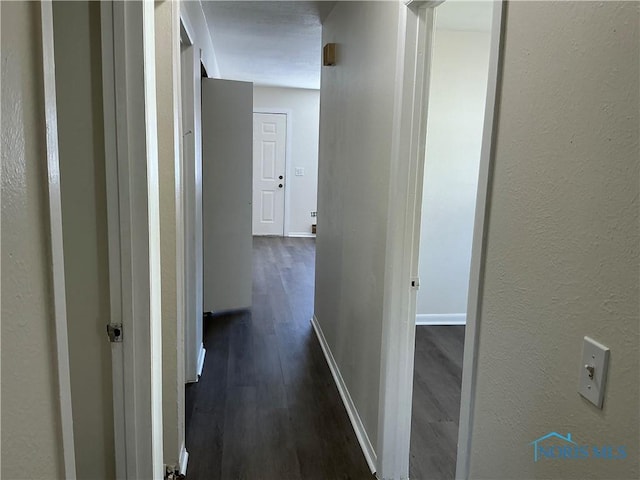 corridor featuring a textured wall, dark wood-type flooring, and baseboards