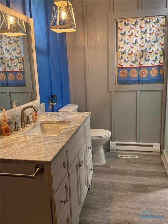 bathroom with toilet, a baseboard radiator, wood finished floors, vanity, and a decorative wall